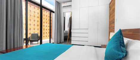 A modern hotel room with a large bed, a view of a brick building, a wardrobe, and a desk with a chair.
