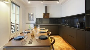 A modern kitchen with a dining area, featuring a wooden table set for four, a built-in stove, and a large window with decorative iron railings. - Sublime Apartment With AC Louvre Museum (Paris)