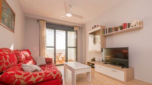 Een woonkamer met een rode bank, een witte salontafel, een televisie op een houten kast en een boekenkast met boeken en decoratieve objecten.