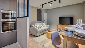 Una cocina moderna con electrodomésticos integrados, un comedor con una mesa lista para dos personas y una sala de estar con sofá y televisor de pantalla plana.