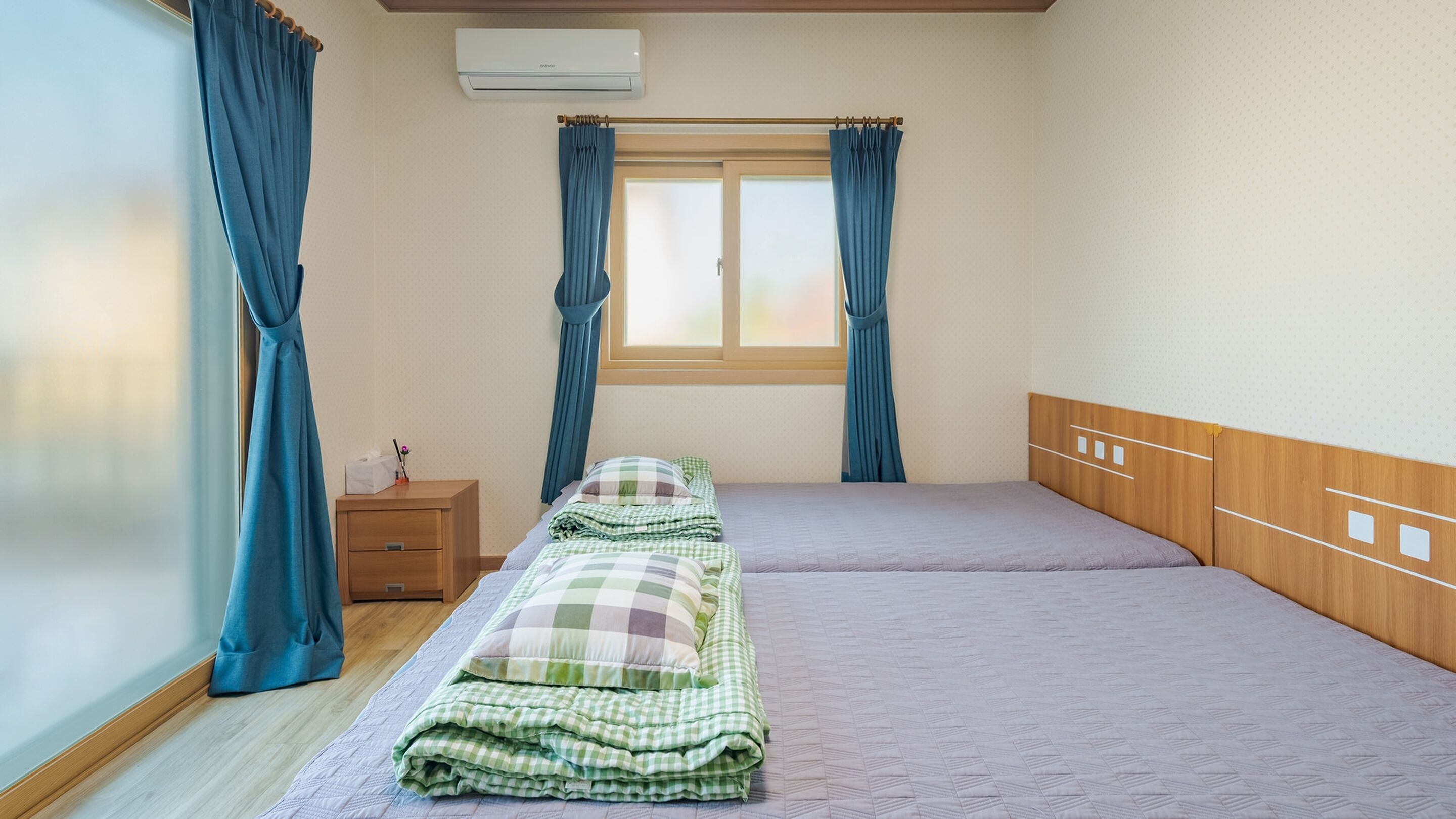 Een eenpersoonskamer met een houten hoofdeinde, een raam met gordijnen, een nachtkastje met een lamp en een wandairconditioner.