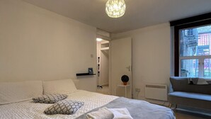 A modern bedroom with a large bed, a chandelier, a window with a view of buildings, and a small table with a plant. - Chamberlayne Inn (London)