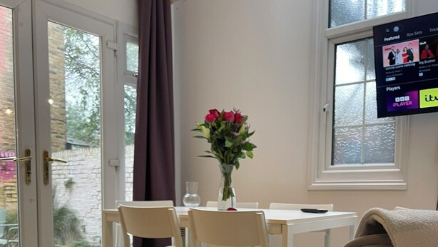 A room with a dining table, chairs, a TV mounted on the wall, and a window with a view of trees. - Chamberlayne Inn (London)
