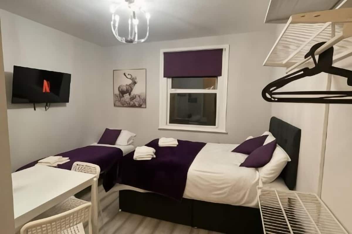 Ein Zimmer mit zwei Betten, einem Schreibtisch, einem Fernseher, einem Fenster mit Vorhang und einer Kleiderstange.