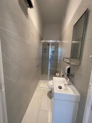 A narrow bathroom with a glass-enclosed shower, a white sink, and a mirror on the wall. - Chamberlayne Inn (London)