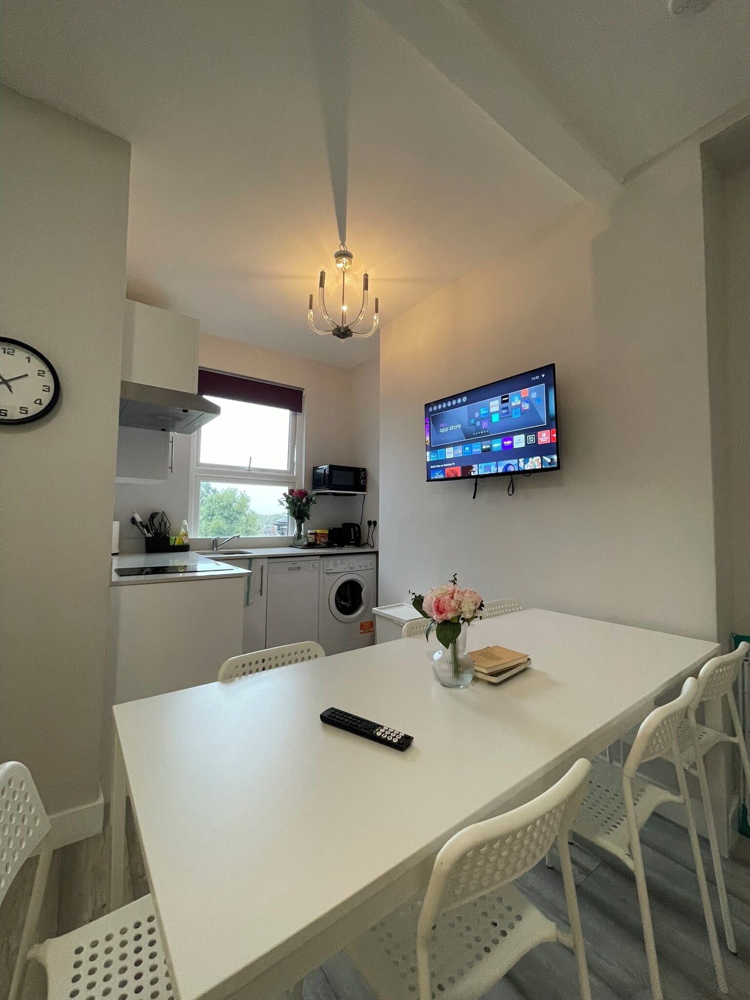 A compact kitchen with a microwave, sink, and a dining area with a table and chairs. A TV is mounted on the wall, and a clock is visible on the wall.