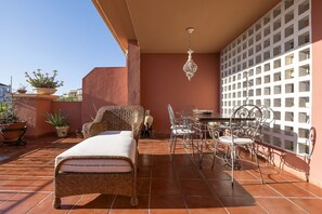 Eine Terrasse mit Liege, Korbmöbeln, einem Esstisch, Stühlen, Topfpflanzen und einer Dekowand.