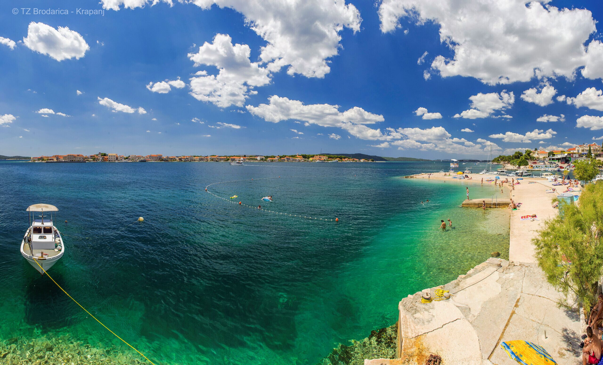 A coastal scene with a boat anchored near the shore, clear turquoise waters, and a beach with people and sun loungers.