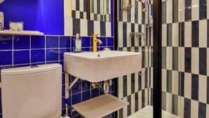 A bathroom with a white toilet, a sink, and a shower. The walls are tiled in blue and black checkered patterns. - WS - Opéra - Milan (Paris)