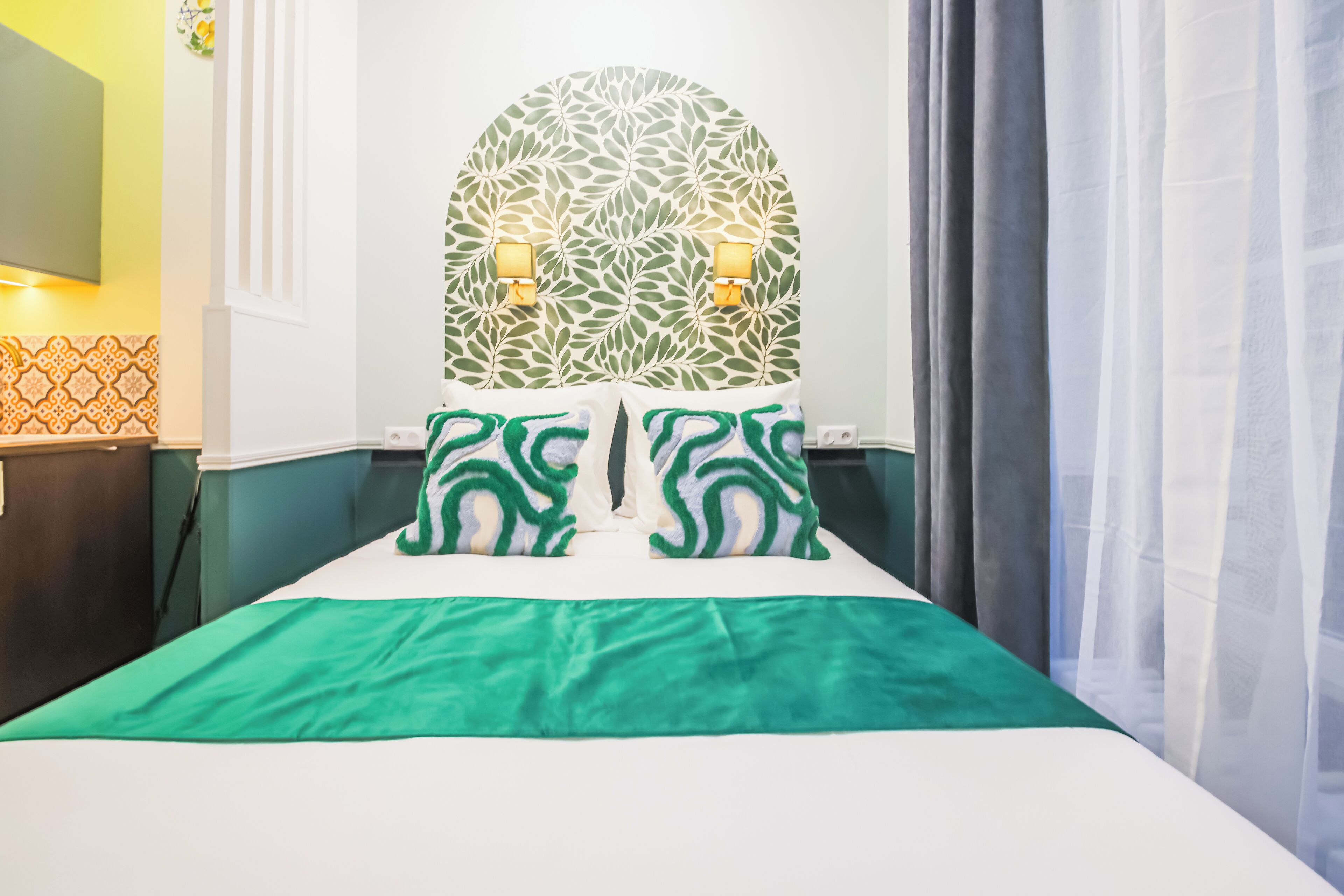Cama bem arrumada com colcha verde e almofadas combinando, luminária fixada na parede e parede com padrão decorativo atrás da cama.