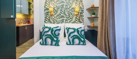 A bedroom with a bed, green and white patterned pillows, a headboard with a leaf pattern, and a view of the kitchen.