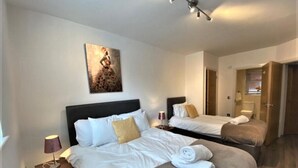 A hotel room with two beds, a nightstand with a lamp, a painting on the wall, and a door leading to another room. - Cosy City Centre Apartment with parking (West Midlands)