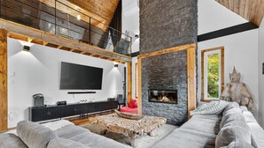 Una sala de estar moderna con un amplio sofá modular, una chimenea de piedra, un televisor montado y una estructura de techo de madera.