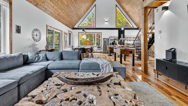 Un espacio habitable amplio con un sofá modular grande, una mesa de centro de madera, y un comedor con techos altos y ventanas grandes.