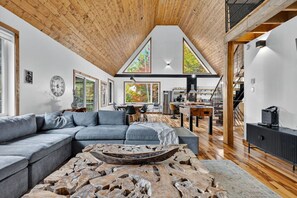 Un espacio habitable amplio con un sofá modular grande, una mesa de centro de madera, y un comedor con techos altos y ventanas grandes.