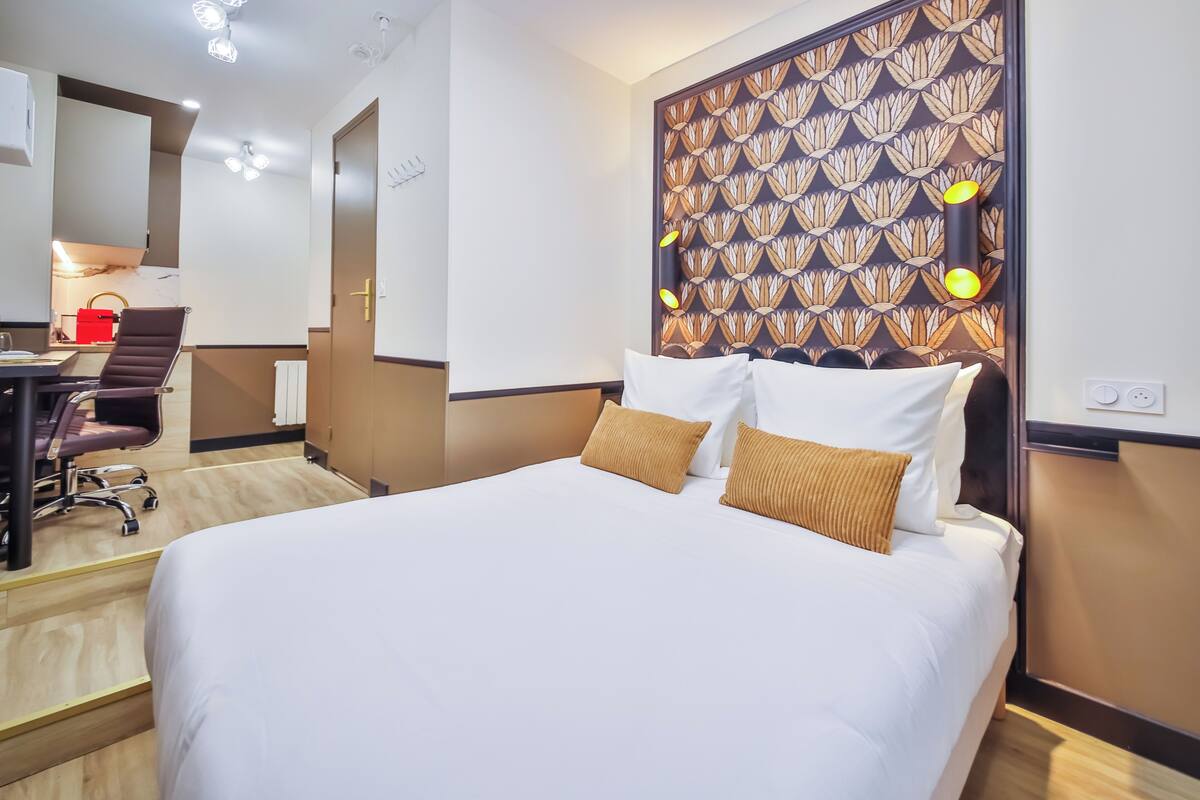 A hotel room with a large bed, a desk with a chair, a red bag, and a patterned wall panel.