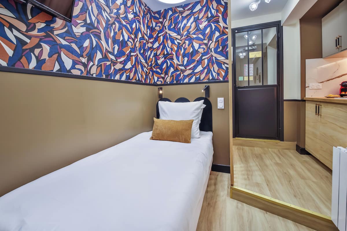 A compact hotel room with a bed, a small kitchenette, and a patterned wall.