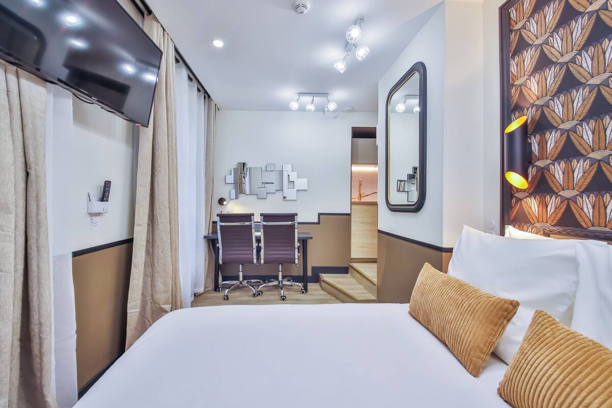 A hotel room with a bed, a desk, two chairs, a mirror, and a patterned wall.