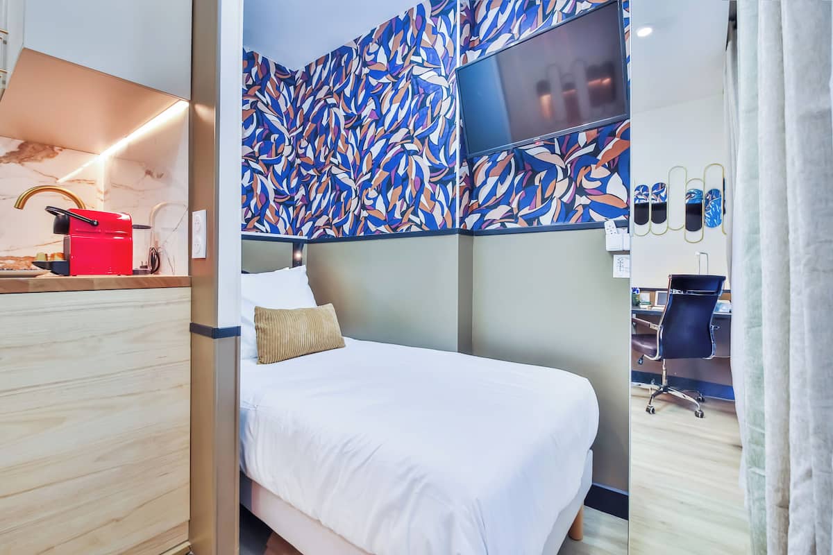 A modern hotel room with a bed, a desk with a red appliance, a large abstract mural, and a TV mounted on the wall.