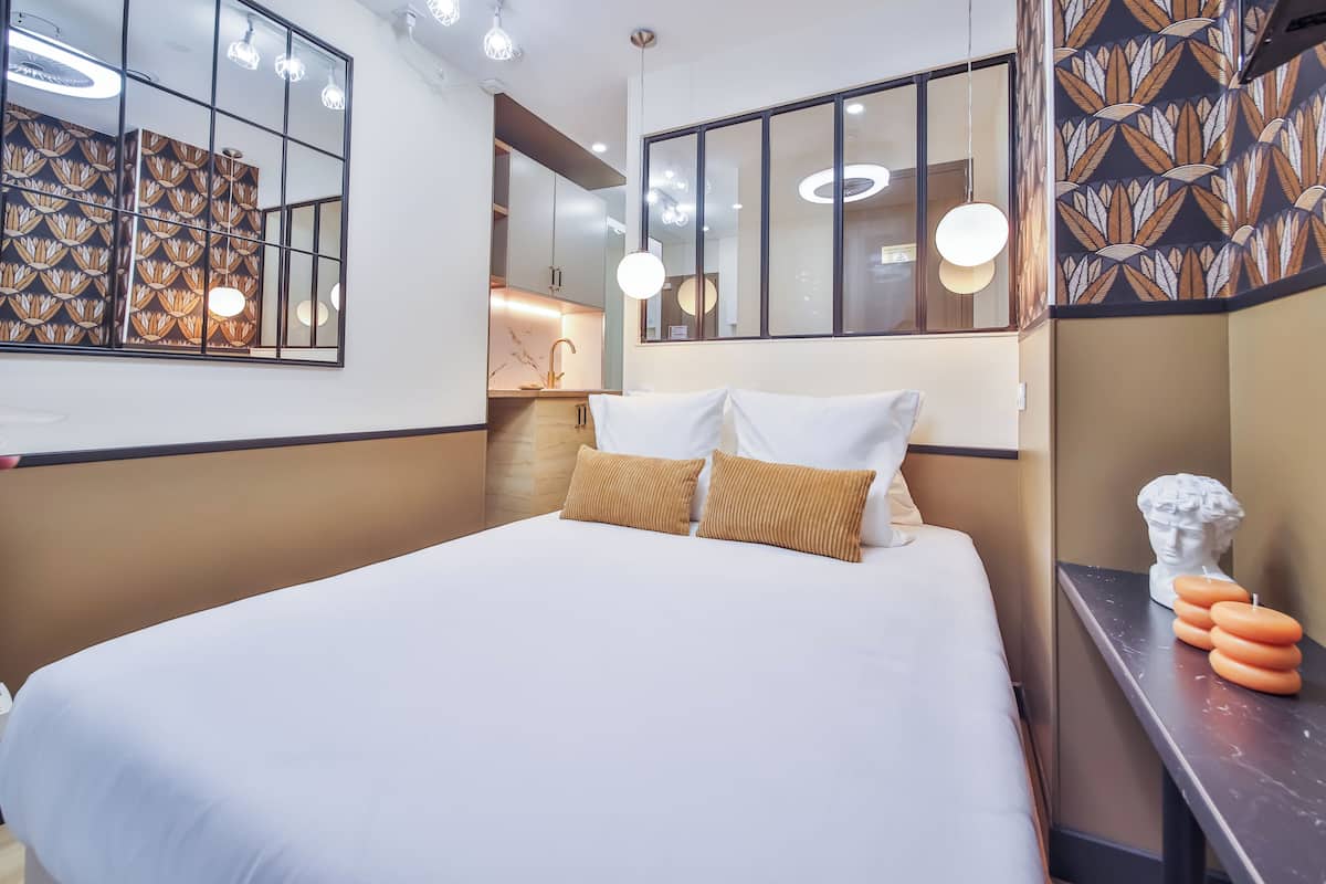 A modern hotel room with a large bed, a bedside table with candles, a mirror, and a decorative wall panel.