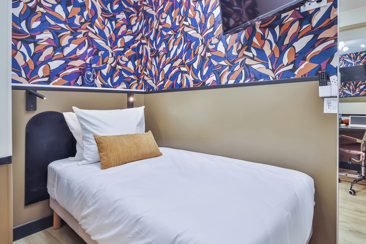 A hotel room with a bed, a headboard, a wall-mounted TV, and a desk with a chair.
