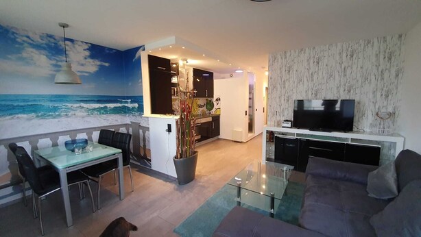 A modern living room with a glass coffee table, a sofa, and a TV. There's a dining area with a table and chairs, and a kitchen with dark cabinets. - Welcome to apartment 20 in house 1 of the Vier Jahreszeiten complex (Großenbrode)