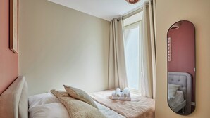 Een slaapkamer met een bed, kussens, een nachtkastje met handdoeken, een raam met gordijnen en een spiegel aan de muur.