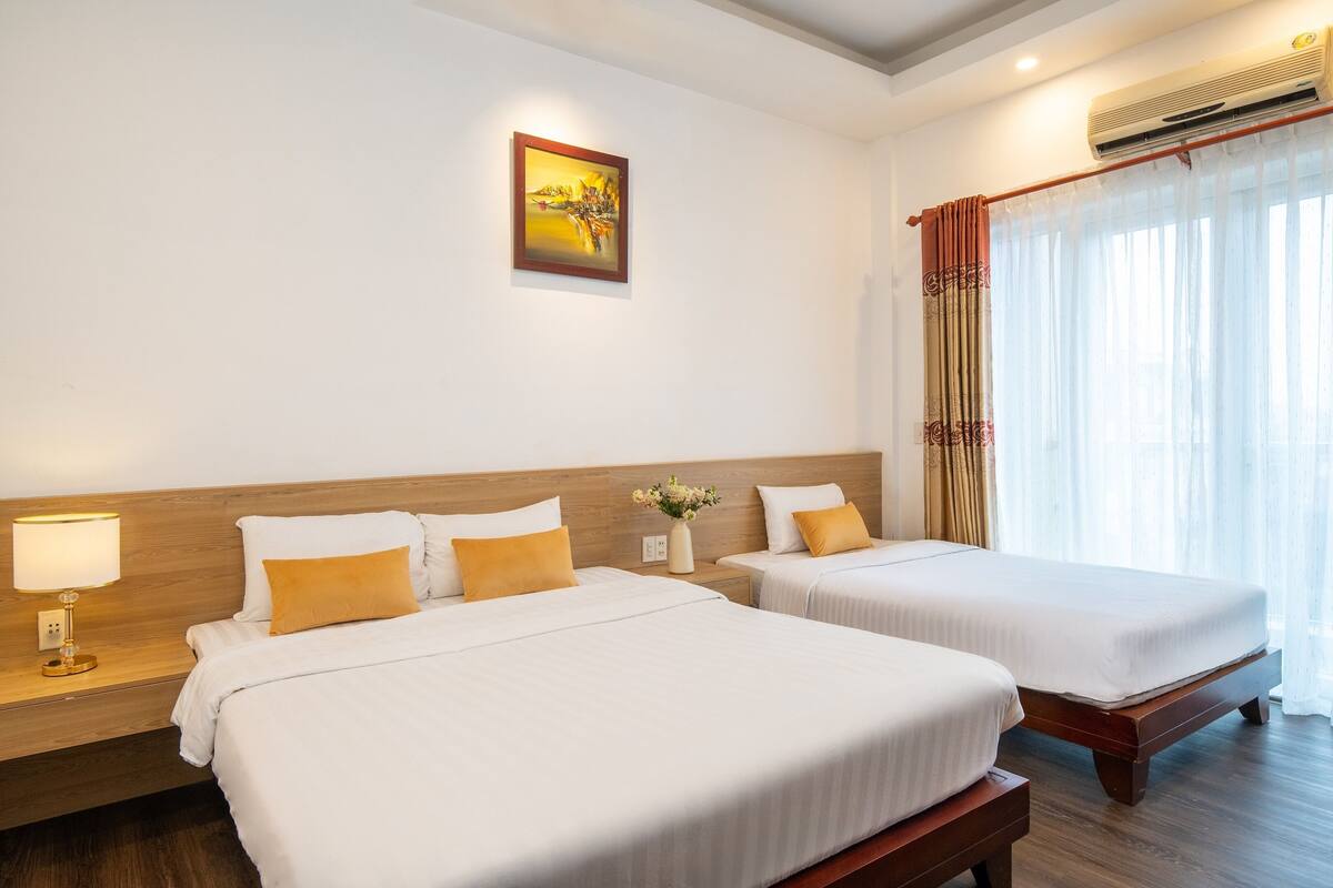 A hotel room with two beds, a nightstand with a lamp, a wall-mounted air conditioner, and a window with sheer curtains.