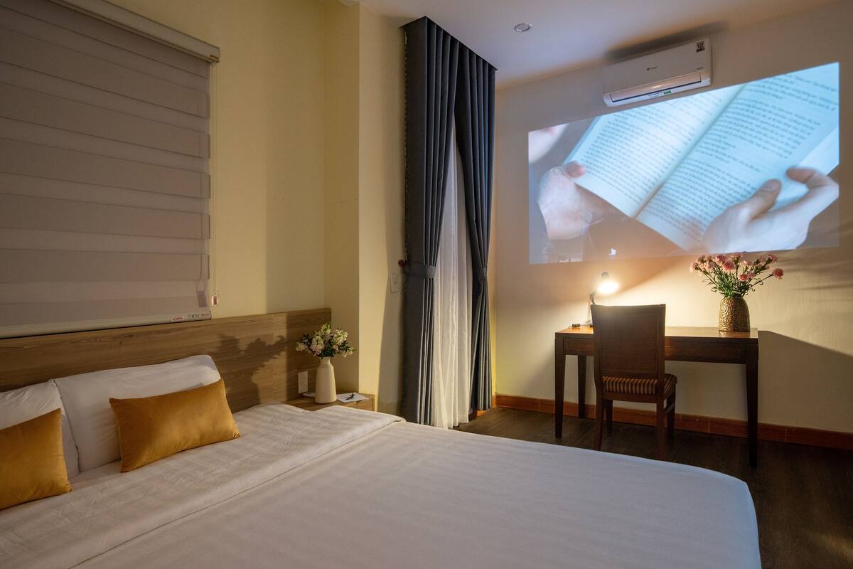 A hotel room with a bed, a desk, a chair, a projector displaying a book, and an air conditioning unit.