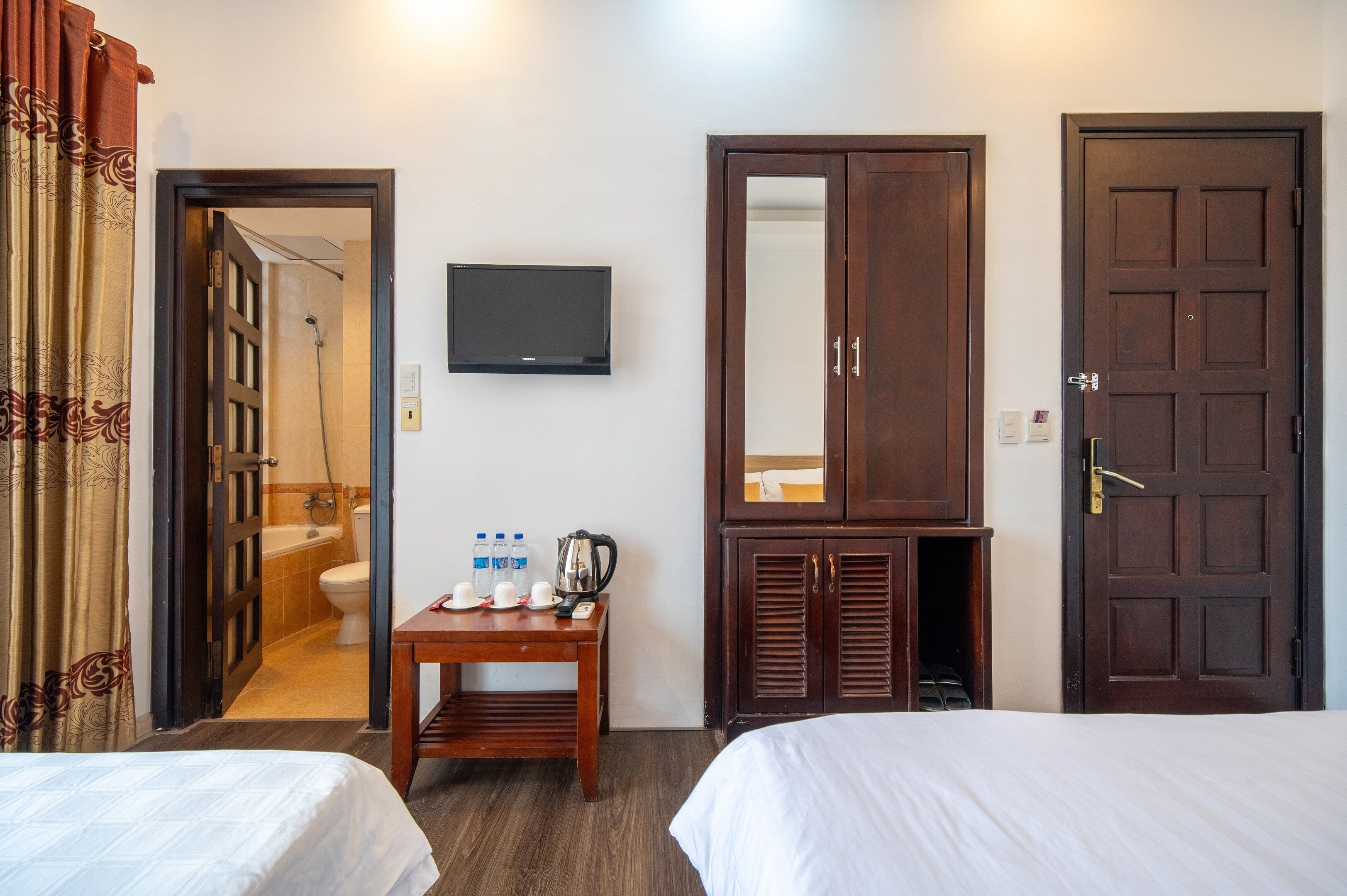 A hotel room with two beds, a wooden cabinet with a mirror, a television on the wall, a small table with toiletries, and a shower area visible through an open door.