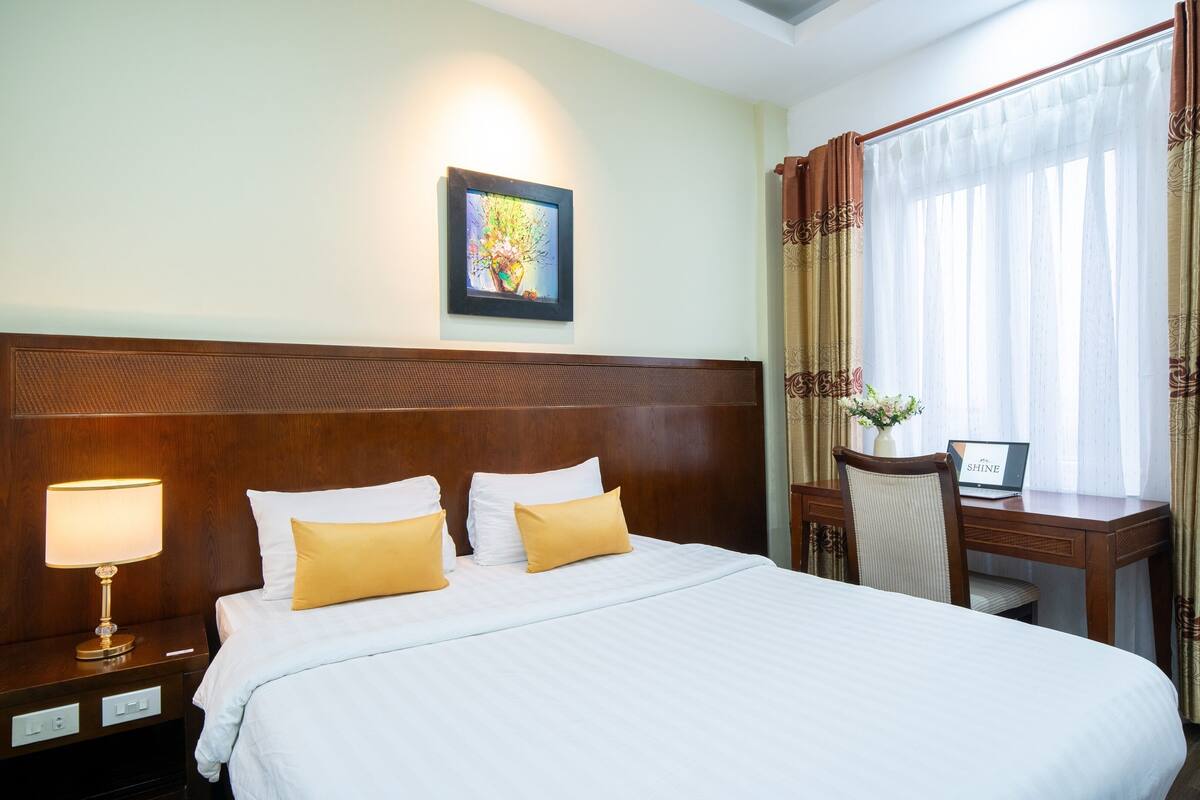 A hotel room with a large bed, a desk with a laptop, a nightstand with a lamp, and a framed picture on the wall.
