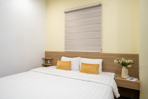 A neatly made bed with white and yellow pillows, a wooden headboard, a bedside table with a vase of flowers, and a window with blinds. - Shine Hue Hotel 2 (Hue)