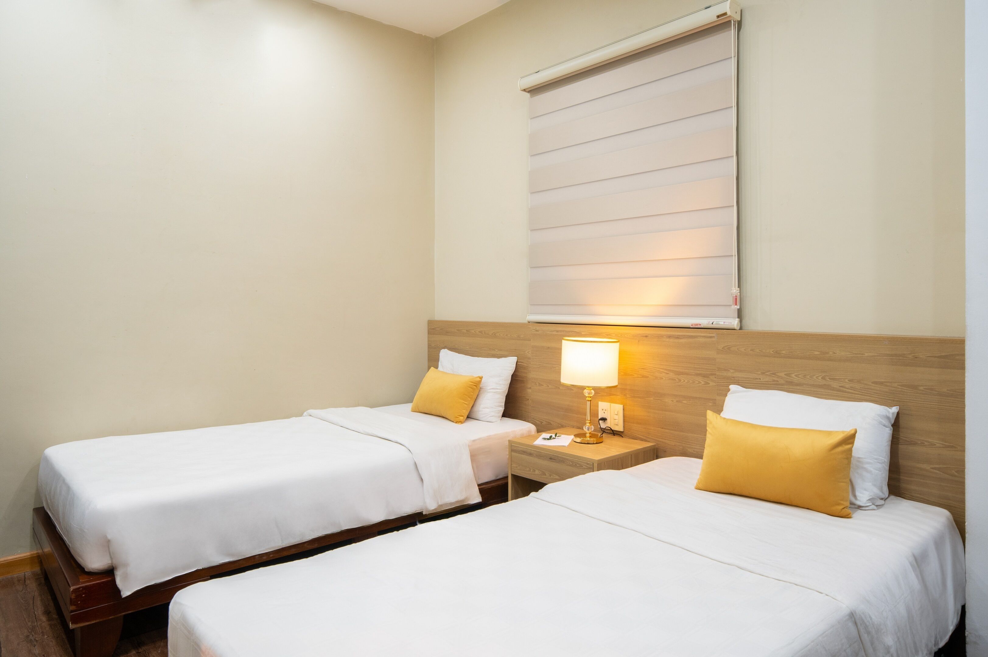 Habitación de hotel con dos camas individuales, cada una con cubrecama blanco y almohada amarilla. Se observa un cabecero de madera, una mesita de noche con lámpara y una ventana con persianas.