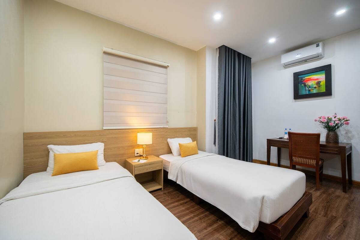 A hotel room with two beds, a desk, a chair, a window with blinds, an air conditioner, and a framed picture on the wall.