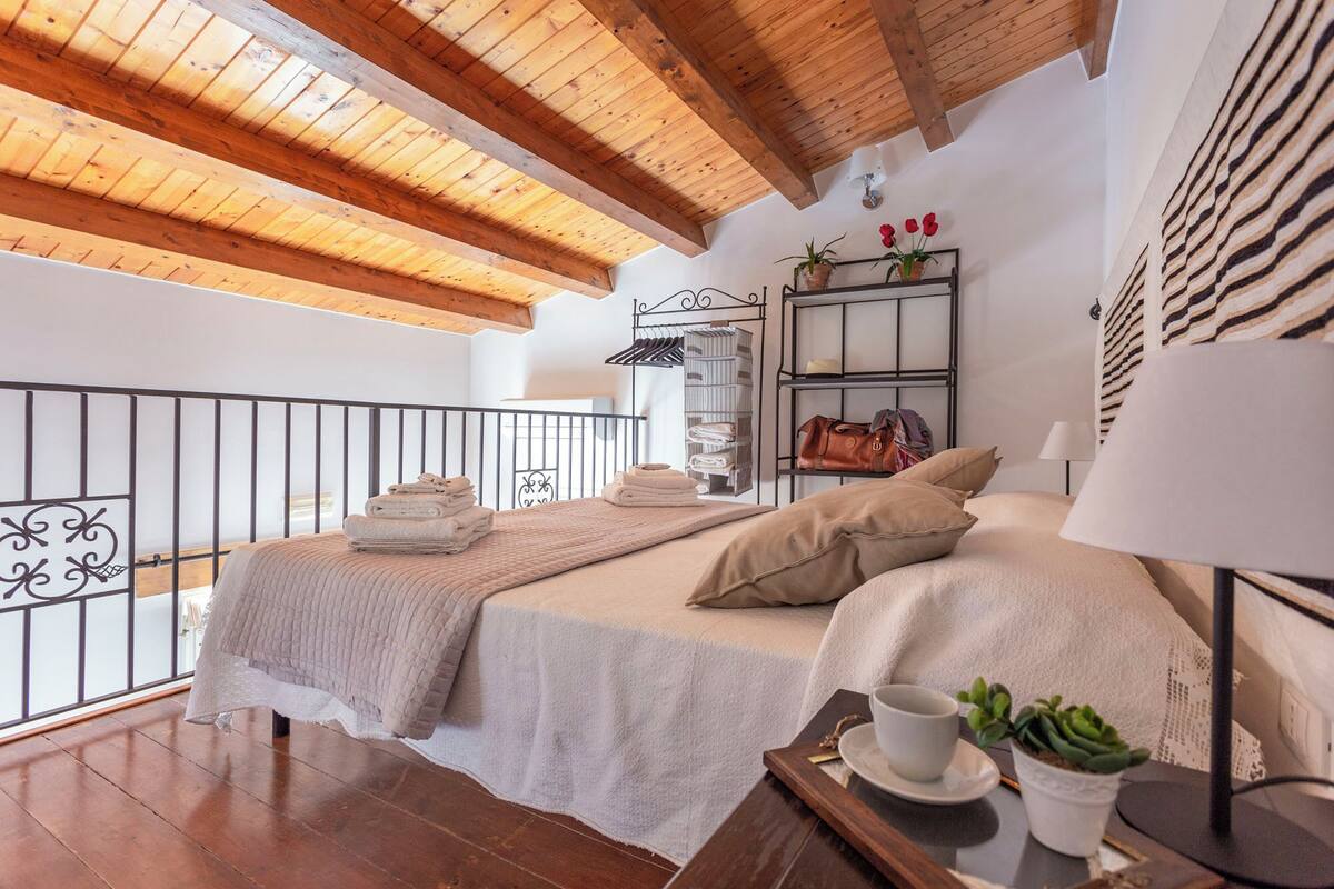A bedroom with a wooden ceiling, a bed with a white bedspread, a bedside table with a cup and a plant, and a metal rack with towels and a handbag.