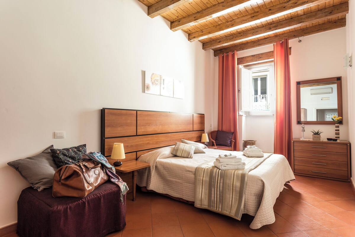 A bedroom with a wooden headboard, a bed with pillows and a blanket, a nightstand with a lamp, a chair, a dresser, and a window with curtains.