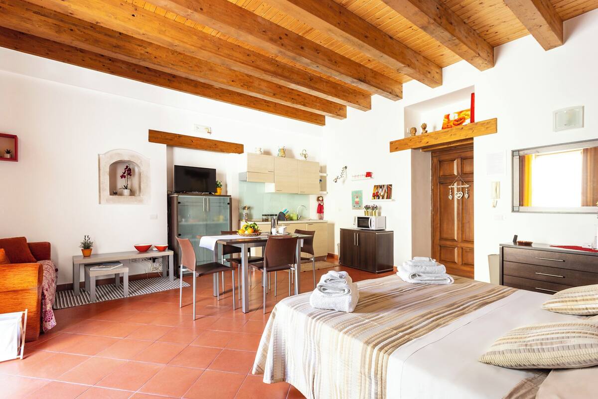 A room with a bed, a dining table, chairs, a kitchen area, and a wooden ceiling.