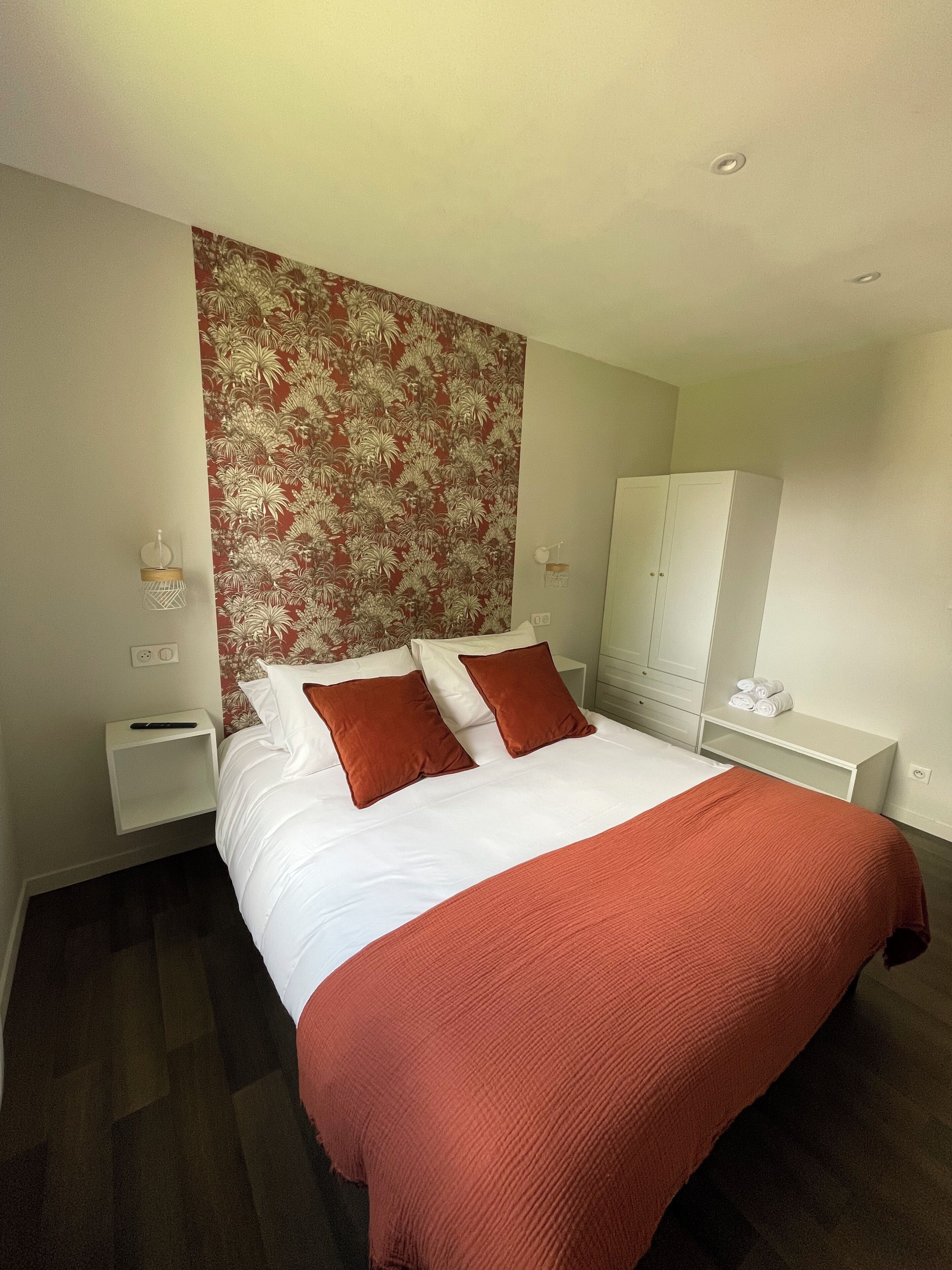 Habitación de hotel con una cama grande, dos almohadas, una colcha roja, una mesita de noche, un armario blanco y una pared con estampado floral.