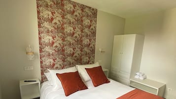 A hotel room with a large bed, two pillows, a red blanket, a nightstand, a white wardrobe, and a floral-patterned wall.