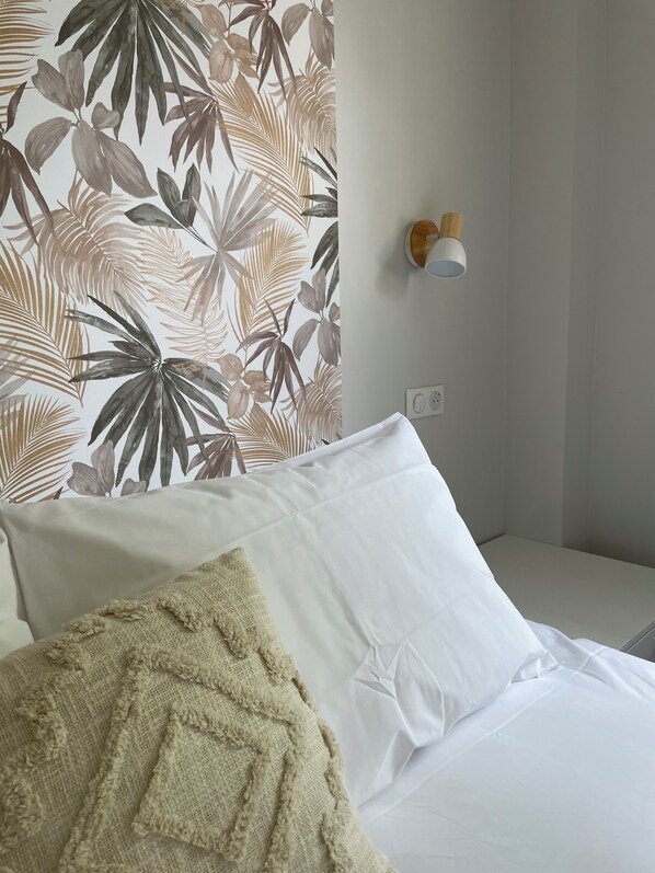 A hotel room with a bed, a wall with tropical leaf wallpaper, a white pillow, and a small wall-mounted light fixture. - Ducey Appart'hôtel (Ducey-les-Chéris)
