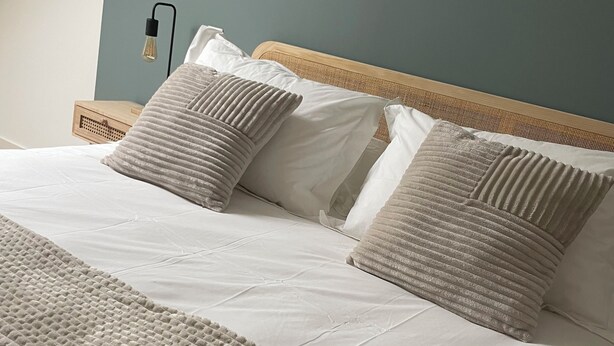 A neatly made bed with a textured blanket and two pillows, a wooden headboard, and a wall-mounted light fixture. - Ducey Appart'hôtel  (Ducey-les-Chéris)