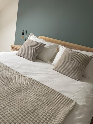 A neatly made bed with a textured blanket and two pillows, a wooden headboard, and a wall-mounted light fixture. - Ducey Appart'hôtel  (Ducey-les-Chéris)