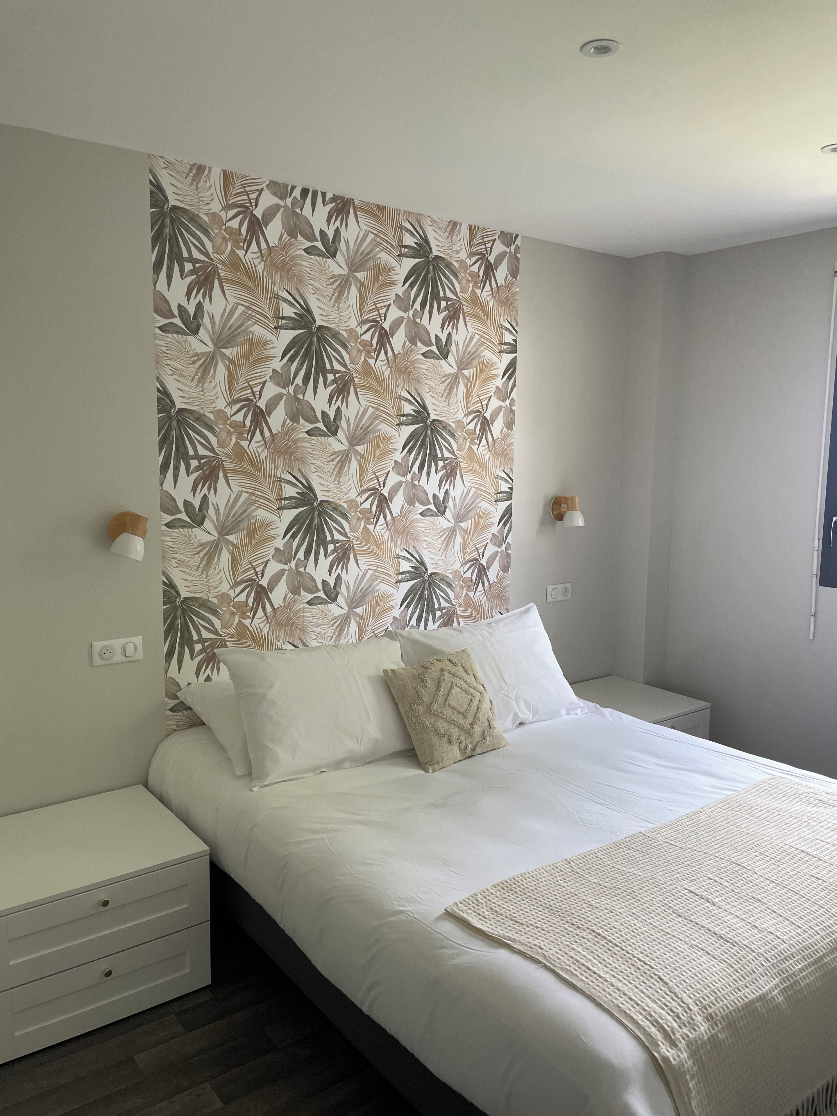Un dormitorio con una cama, dos mesitas de noche, una pared con estampado de hojas tropicales y dos lámparas de pared.