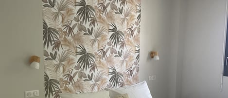 Une chambre à coucher comprenant un lit, deux tables de chevet, un mur orné d’un motif de feuilles tropicales et deux lampes fixées au mur.