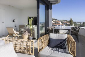 A balcony with wicker furniture, a dining table, and a view of a coastal town.