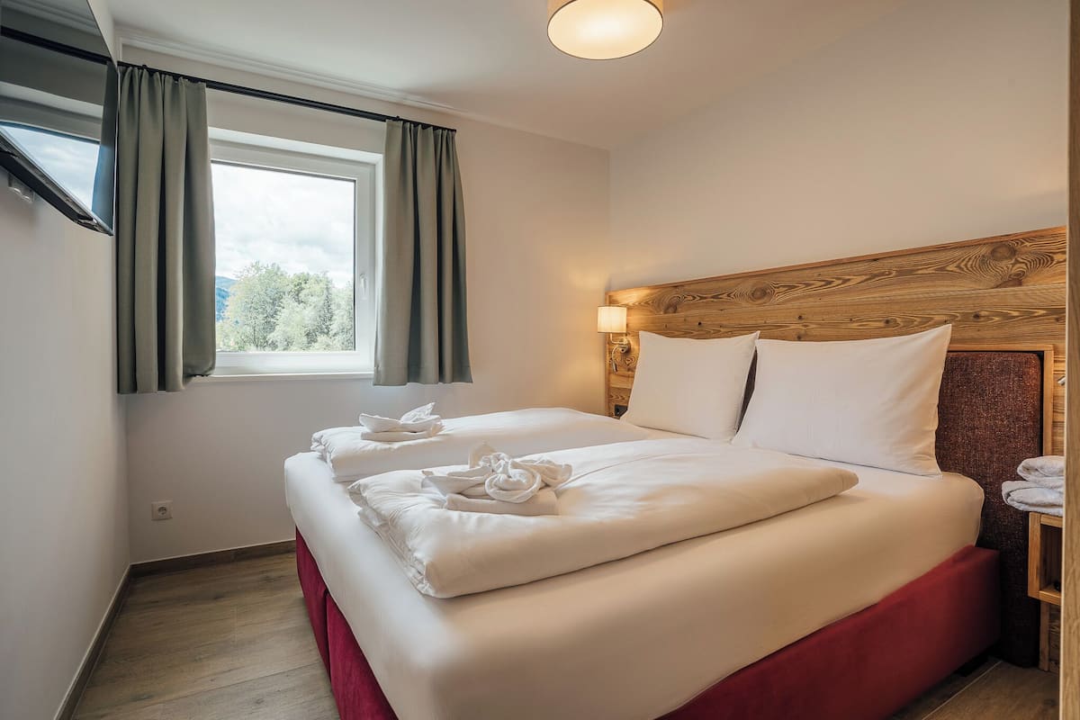 Habitación de hotel con dos camas, cabecera de madera, ventana con cortinas y mesita de noche con toallas.