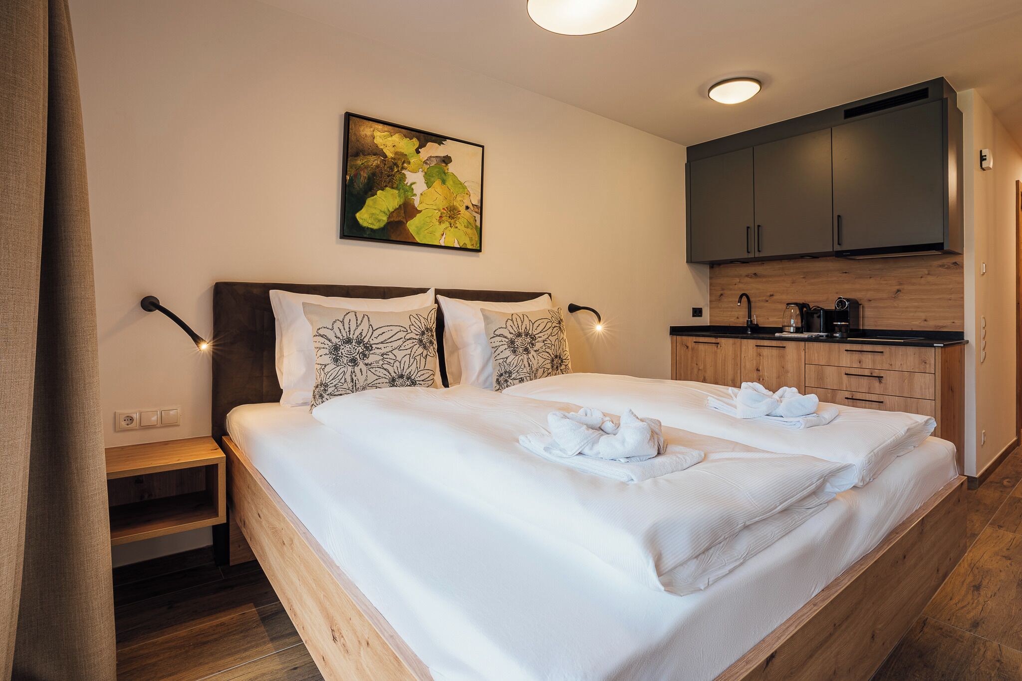 Ein Schlafzimmer mit einem großen Bett, zwei Kissen, einem Nachttisch, einem hölzernen Kopfteil, einem Bild an der Wand und einer Küchenzeile mit Schränken und einer Arbeitsfläche.