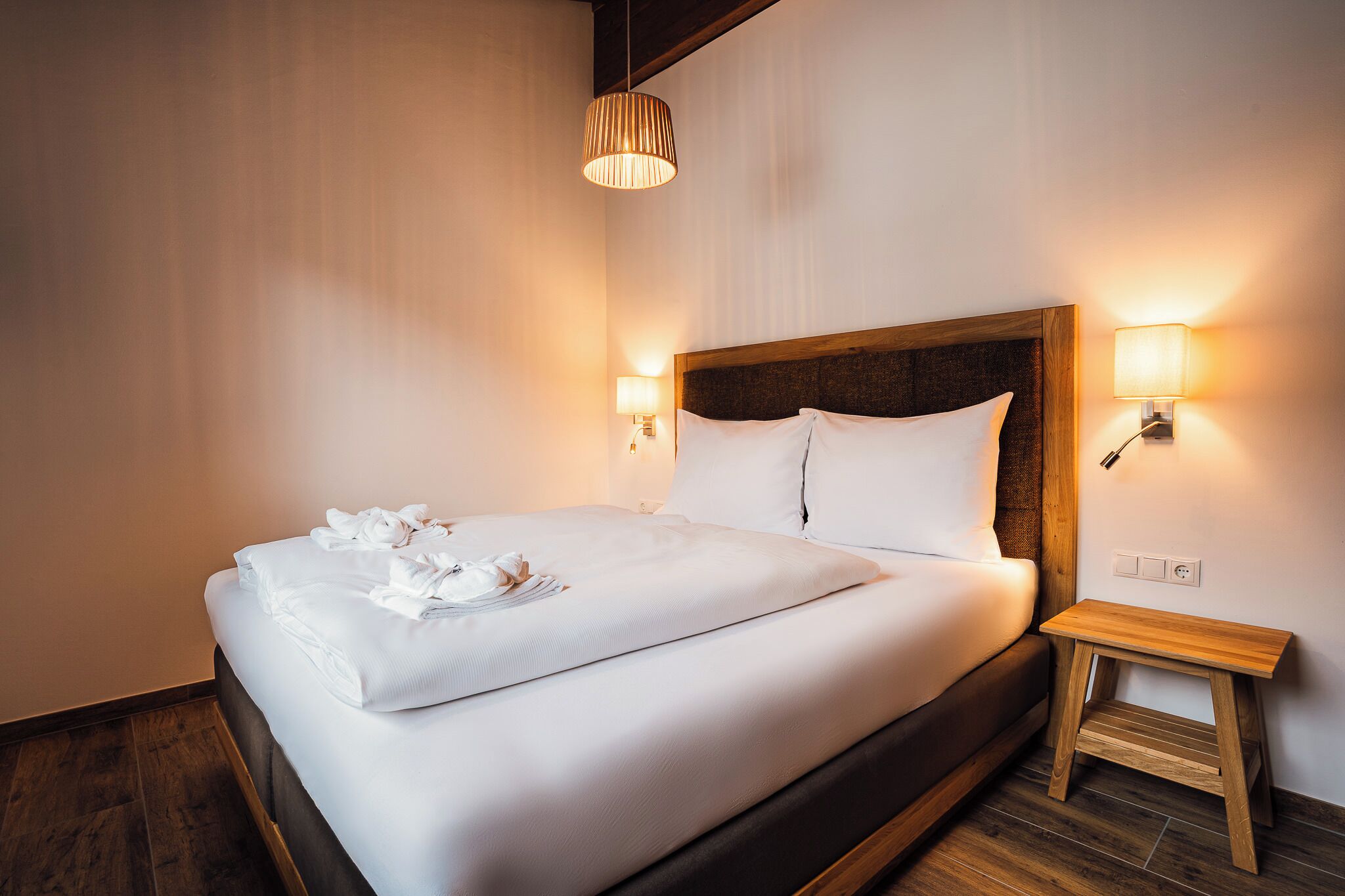 Un lit bien fait, avec du linge de lit et des serviettes blanches, une tête de lit en bois et deux lampes de chevet.