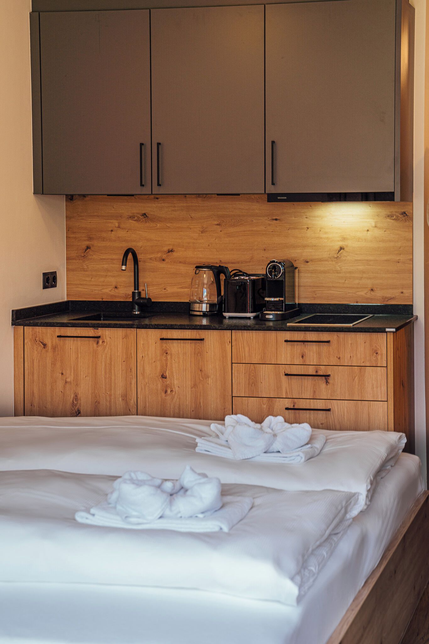 Une chambre d’hôtel compacte avec un coin cuisine en bois, un lit agrémenté de serviettes blanches soigneusement pliées et une grande fenêtre laissant entrer la lumière naturelle.