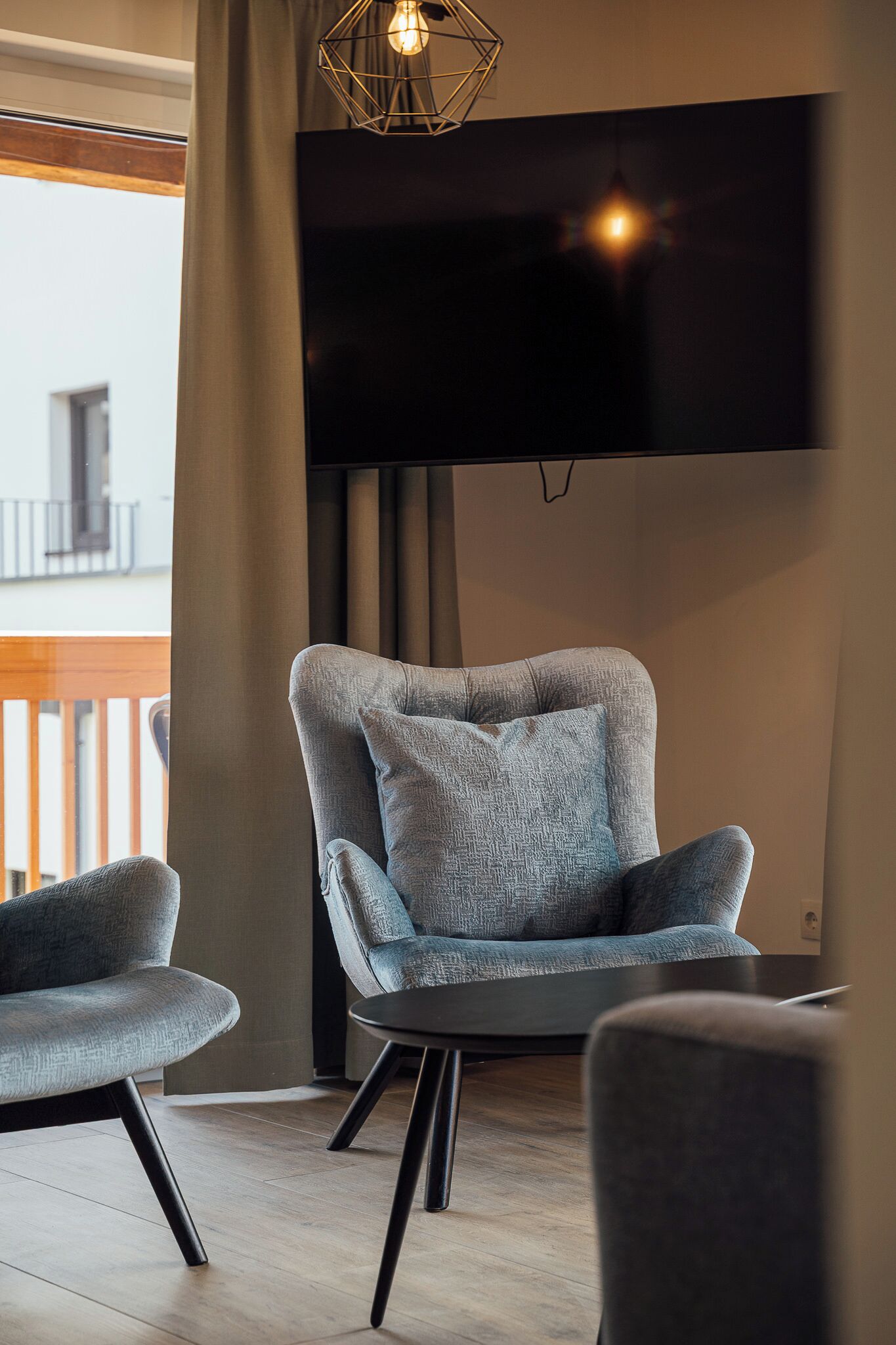 A modern living room with a grey upholstered armchair, a smaller armchair, and a black coffee table. A flat-screen TV is mounted on the wall above the armchairs.